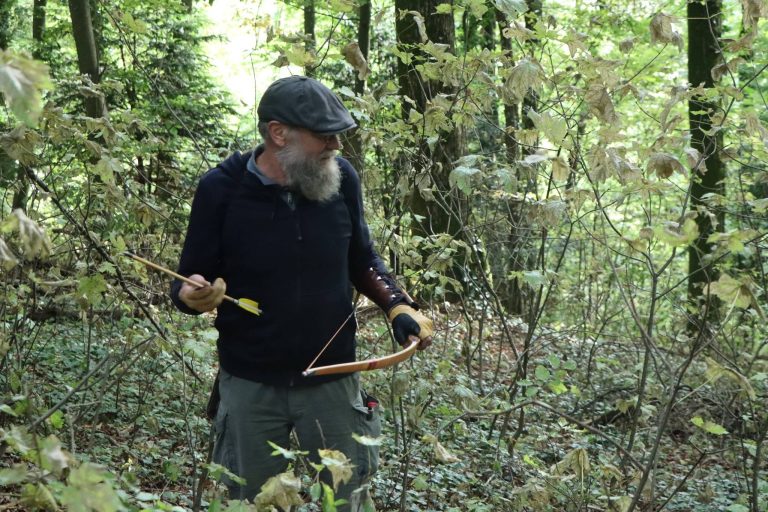 Bärtiger Bogenschütze mit Mütze hält Pfeil und Bogen im Wald
