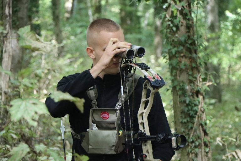Junger Bogenschütze mit Compoundbogen schaut im Wald durch ein Fernglas