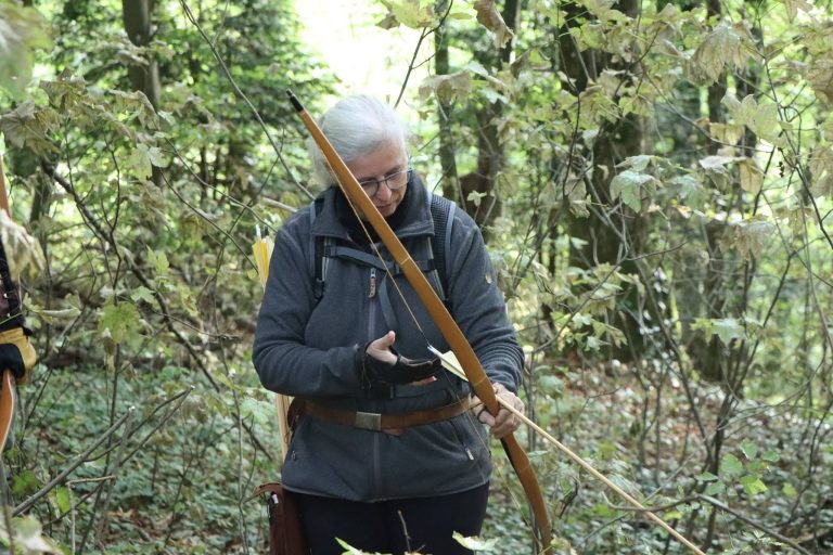 Grauhaarige Bogenschützin in grauer Jacke hält ihren Langbogen im Wald