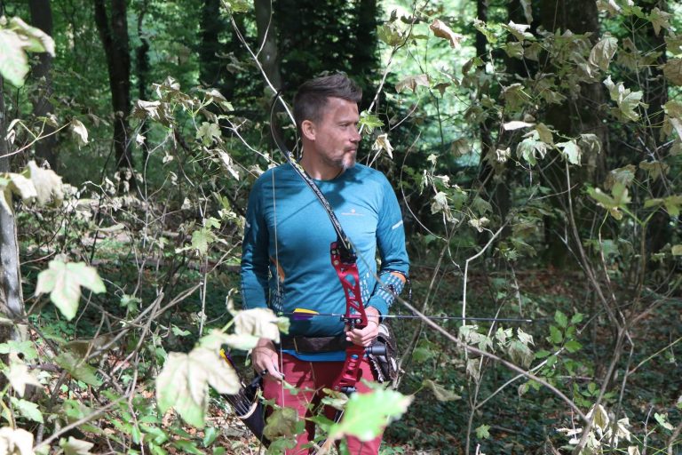 Mann in blauer Jacke steht mit seinem Bogen zwischen Blättern im Wald
