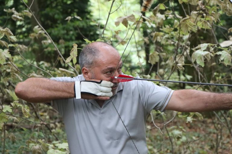 Mann in grauem Shirt spannt den Bogen mit einem roten Pfeil im Wald