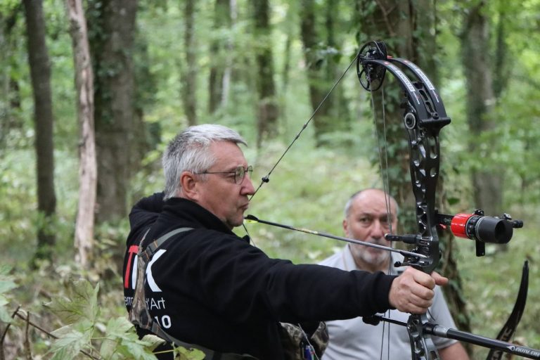 Bogenschütze mit Brille zielt mit einem schwarzen Compoundbogen, dahinter ein weiterer Schütze