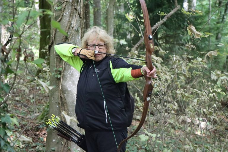 Blonde Bogenschützin in schwarz-grüner Jacke spannt einen Recurvebogen im Wald