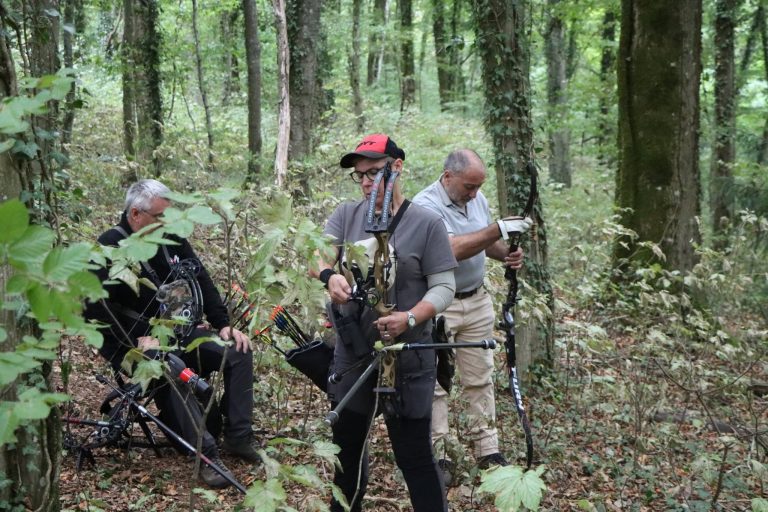 Drei Bogenschützen warten im Wald an einer Station, einer sitzt auf einem Hocker