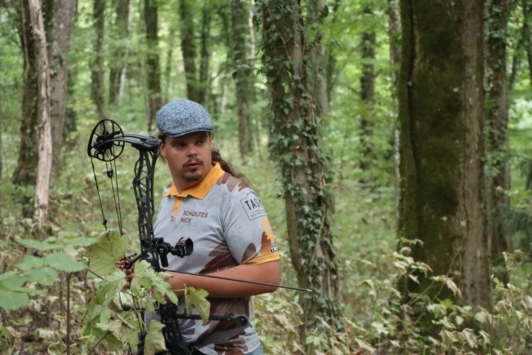 Bogenschütze mit Schiebermütze hält einen Compoundbogen im Wald