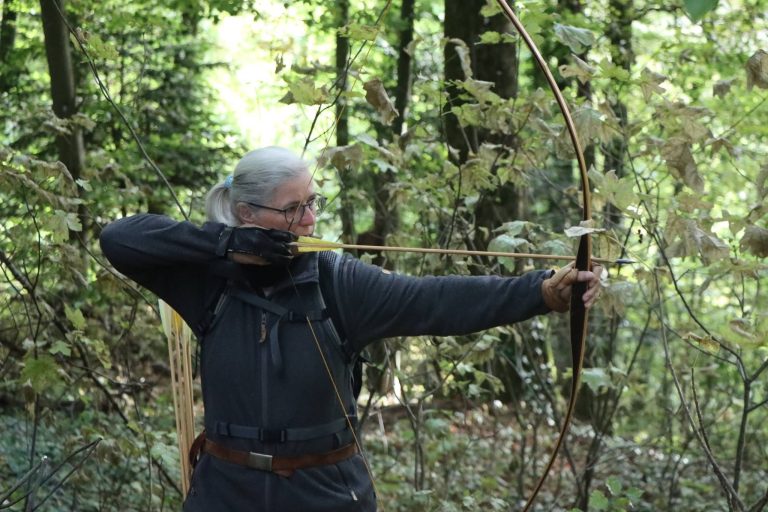 Grauhaarige Bogenschützin mit Brille spannt einen Langbogen im Wald