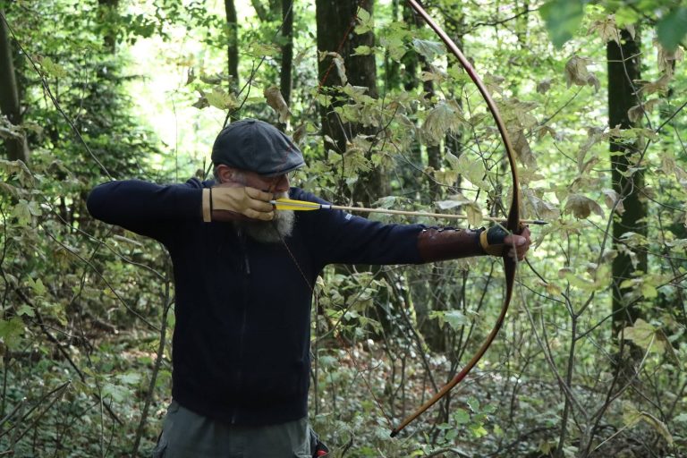 Bogenschütze mit Schiebermütze spannt einen Langbogen im Wald