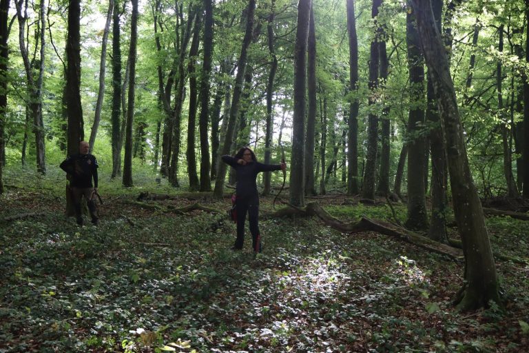 Zwei Bogenschützen zwischen hohen Bäumen in einem schattigen Wald