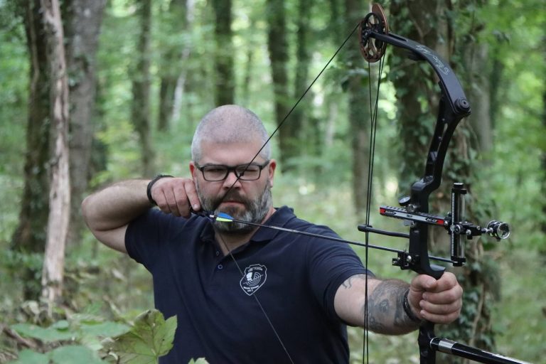 Bärtiger Bogenschütze mit Brille zielt mit einem Compoundbogen im Wald