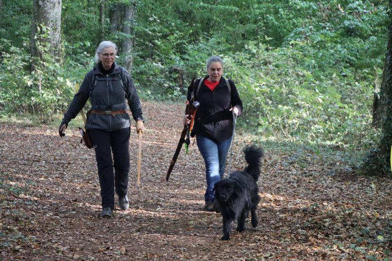 Zwei Frauen gehen mit ihren Bögen und einem schwarzen Hund über einen Waldweg