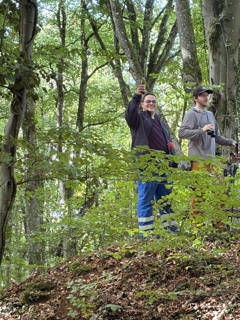 Zwei Männer stehen auf einem Erdhügel im Wald, einer fotografiert mit dem Handy