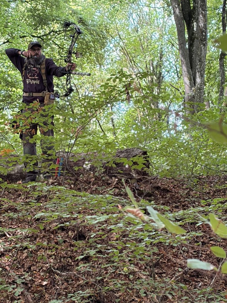 Bogenschütze mit Compoundbogen steht auf einem bewachsenen Hang im Wald