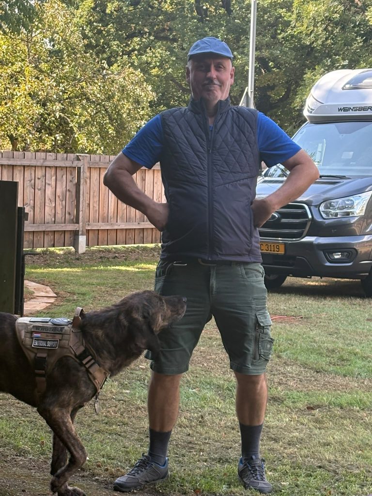 Mann in blauer Weste steht mit seinem Schäferhund auf der Wiese