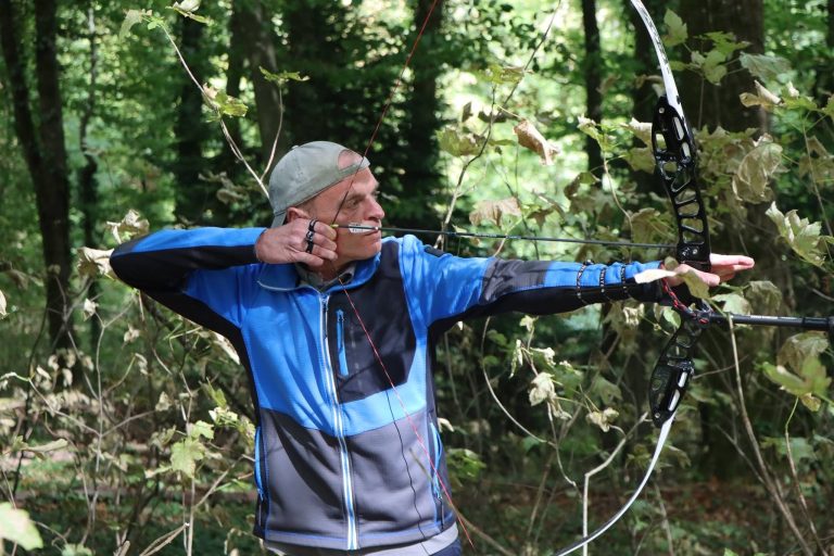 Bogenschütze mit Mütze und blauer Jacke spannt einen Compoundbogen im Wald