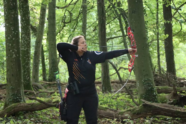 Bogenschützin in schwarzer Kleidung spannt einen roten Recurvebogen im Wald