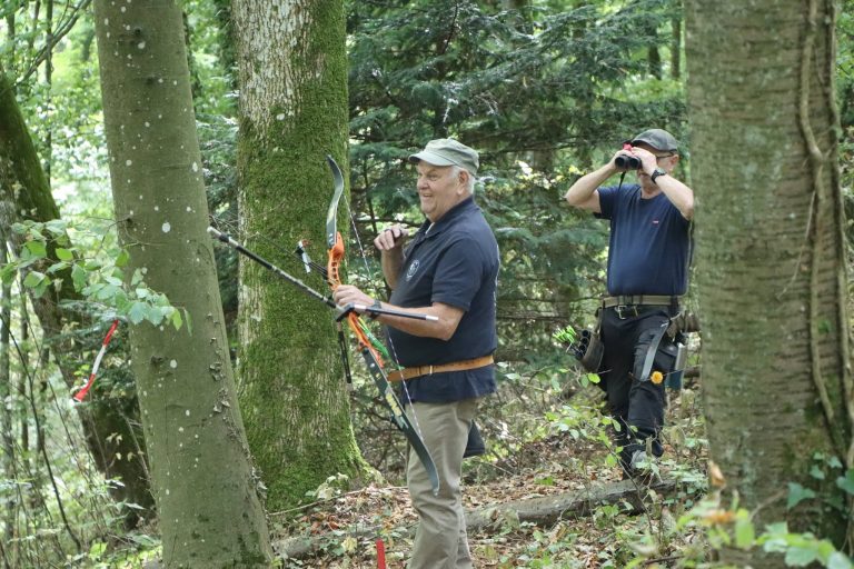 Bogenschütze mit Recurvebogen im Wald, daneben ein Mann mit Fernglas