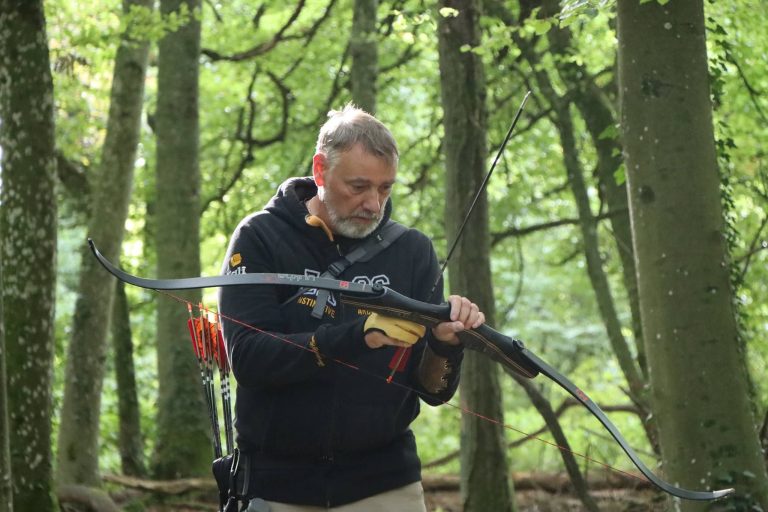 Grauhaariger Bogenschütze mit Bart hält seinen Recurvebogen im Wald