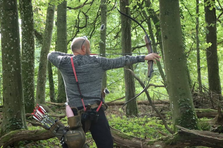 Bogenschütze mit rotem Hosenträger spannt einen Langbogen im Wald, von hinten gesehen