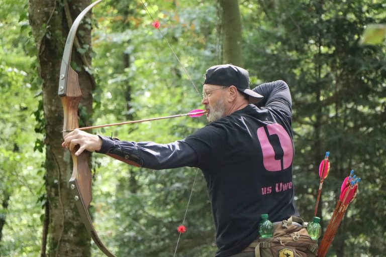 Bogenschütze mit Kappe und Seitenköcher spannt einen Langbogen im Wald