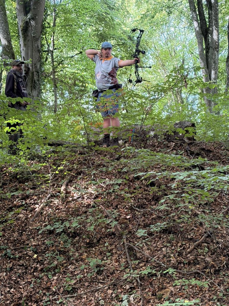 Bogenschütze zielt mit einem Compoundbogen von einer Anhöhe im Wald