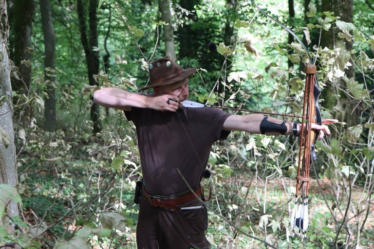 Bogenschütze mit Hut und brauner Kleidung spannt einen Langbogen im Wald