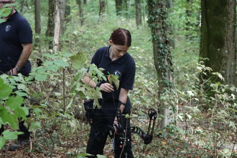 Junge Bogenschützin mit Compoundbogen steht zwischen Pflanzen im Wald