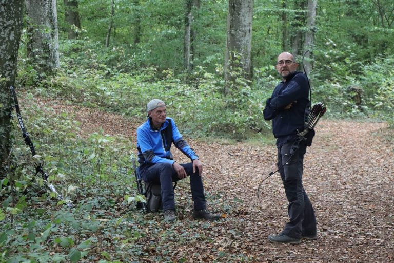 Bogenschütze sitzt auf einem Hocker im Wald, ein anderer steht mit seinem Bogen daneben
