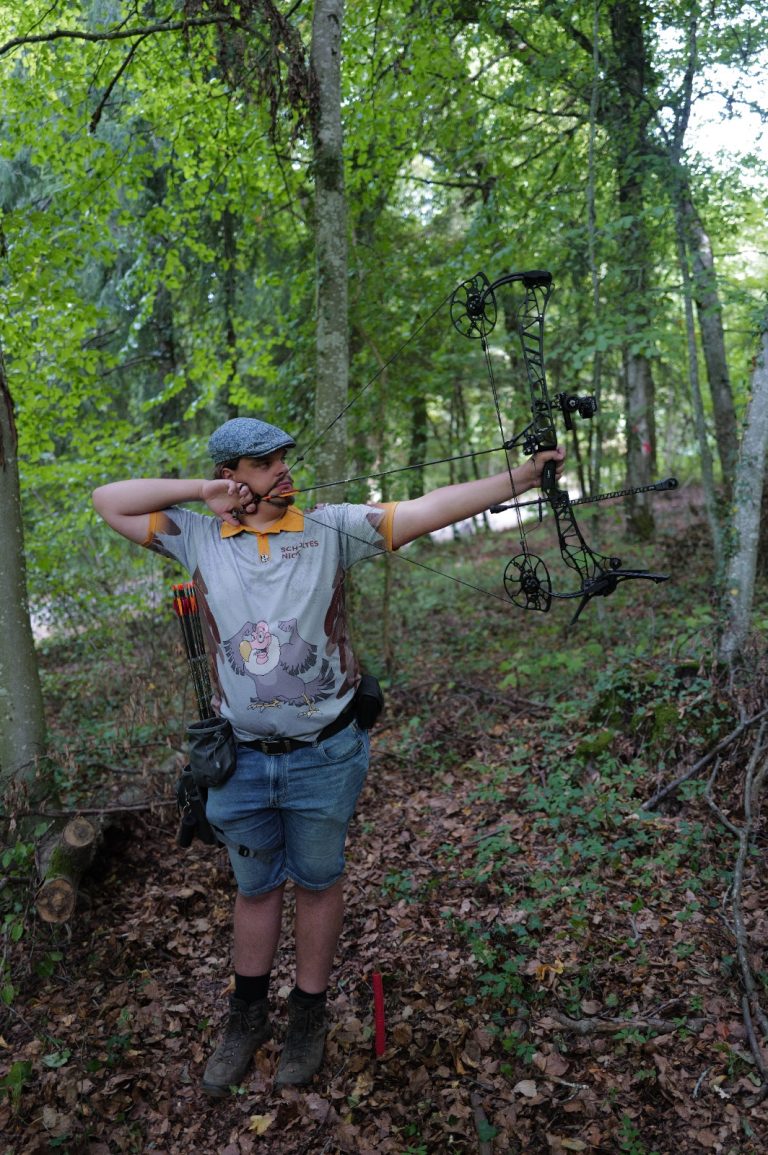 Bogenschütze mit Schirmmütze und Jeansshorts zielt mit dem Compoundbogen im Wald