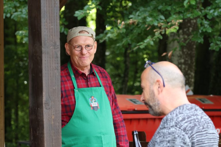 Lächelnder Mann mit grüner Schürze und Kappe am Grill im Gespräch mit einem Gast