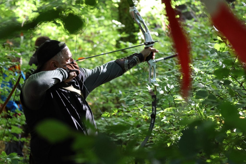 Person mit Stirnband spannt einen Recurvebogen im Wald, rot-weißes Absperrband im Vordergrund