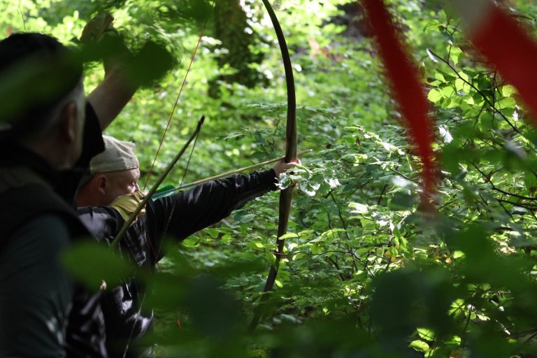 Bogenschütze mit Holzbogen zielt im Wald, ein weiterer Teilnehmer steht daneben