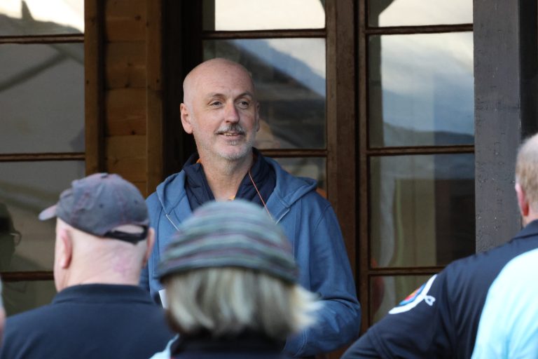 Lächelnder Mann in blauer Jacke spricht vor dem Vereinshaus zu einer Gruppe von Bogenschützen