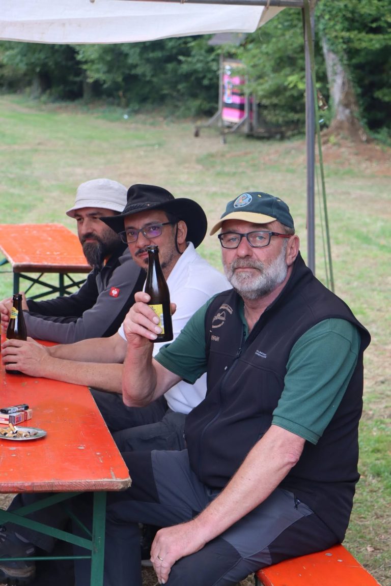 Drei Männer mit Hüten und Kappen sitzen lachend am Biertisch auf der Wiese