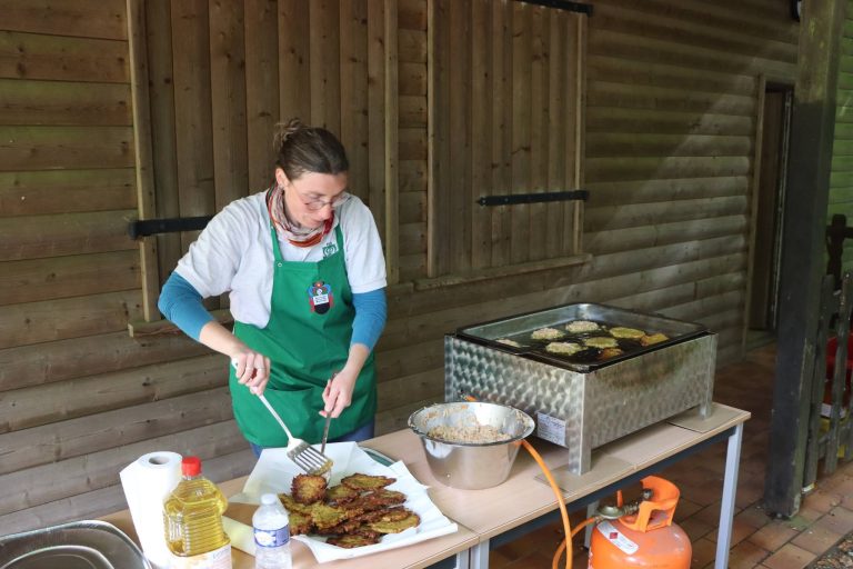 Frau mit Mütze und grüner Schürze grillt unter dem Vordach des Vereinshauses