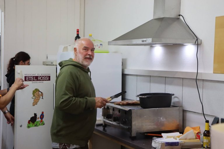 Grauhaariger Mann in grüner Jacke steht lächelnd am Herd mit Pfanne und Topf