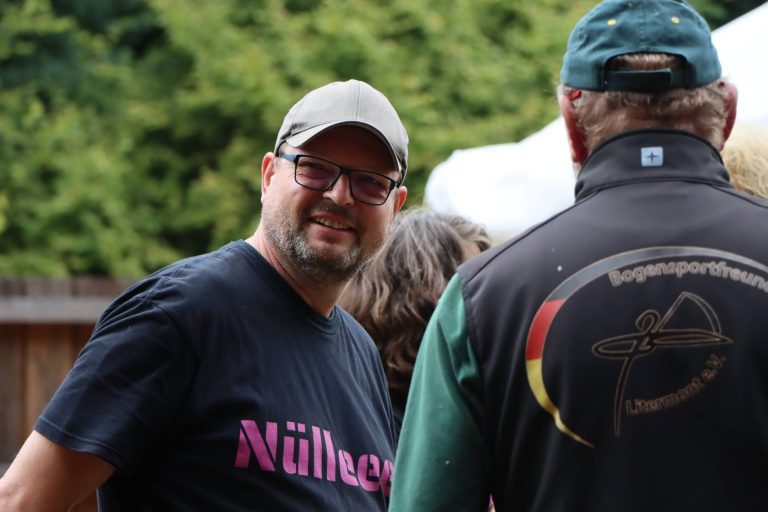 Mann mit Brille und grauer Kappe lächelt in die Kamera, daneben ein Schütze mit Vereinsjacke