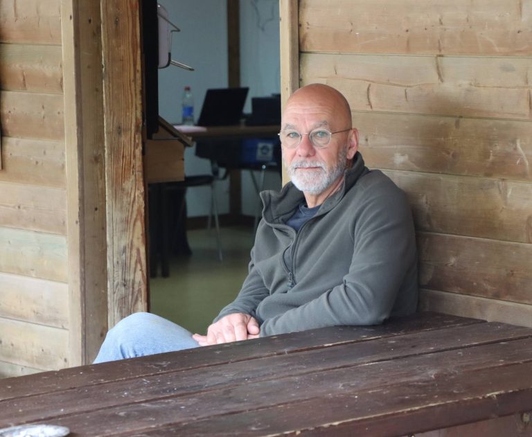 Mann mit Brille in grauem Pullover sitzt entspannt am Holztisch unter dem Vordach