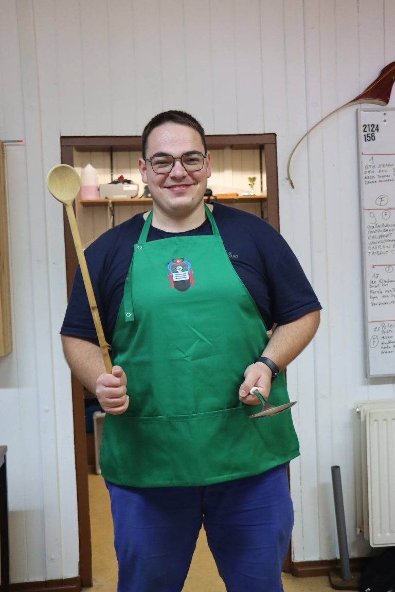 Junger Mann mit grüner Schürze hält einen großen Holzlöffel im Vereinshaus