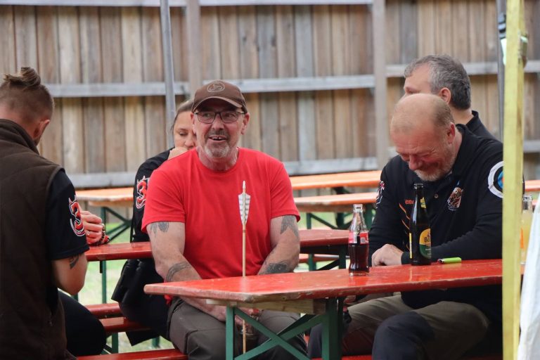 Mann im roten T-Shirt mit Kappe lacht am Tisch unter dem Zelt