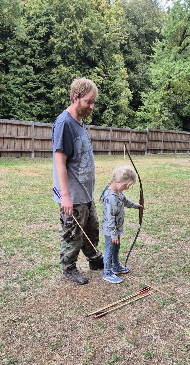 Erwachsener zeigt einem kleinen Kind auf der Wiese, wie man einen Bogen hält